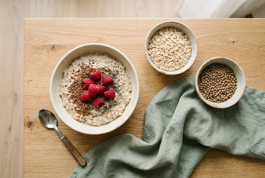 Schüssel Porridge mit Himbeeren, Leinsamen und Chiasamen neben Schalen mit Haferflocken und Linsen auf einem Holztisch